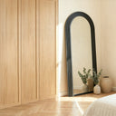 Espejo BADEN de arco apoyado en la pared de un dormitorio, junto a un armario de madera