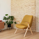 Butaca DINS con patas de haya natural, en mostaza, junto a una pared de ladrillo – Teulat
