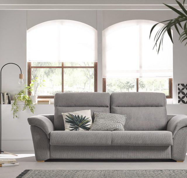 Sofá gris moderno con cojines decorativos y ventanas grandes.