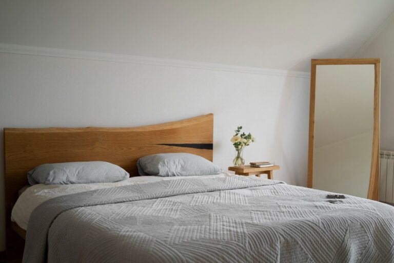 Vista de una cama en una habitación con un espejo de cuerpo y marco de madera al lado y una mesita.